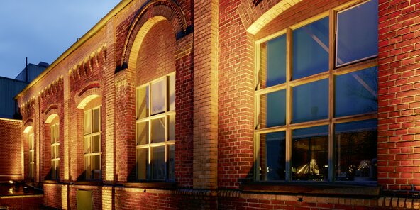 Ziegelsteinfassade eines Gebäudes mit großen Fenstern, die von warmem Licht beleuchtet werden. Die Architektur zeigt Bögen über den Fenstern und eine ruhige Abendstimmung.