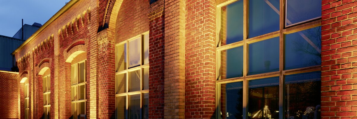 Ziegelsteinfassade eines Gebäudes mit großen Fenstern, die von warmem Licht beleuchtet werden. Die Architektur zeigt Bögen über den Fenstern und eine ruhige Abendstimmung.