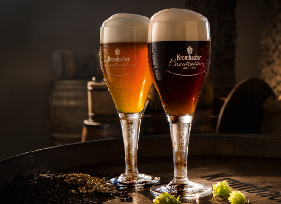 Zwei Gläser Krombacher Bier stehen auf einem Holzfass. Das linke Glas enthält Kellerbier, das rechte dunkles Bier.