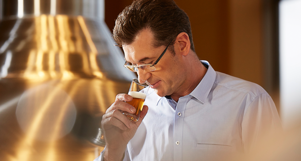 Ein Mann in einem hellblauen Hemd hält ein Glas mit einem goldenen Getränk. Im Hintergrund ist ein metallischer Braukessel sichtbar.