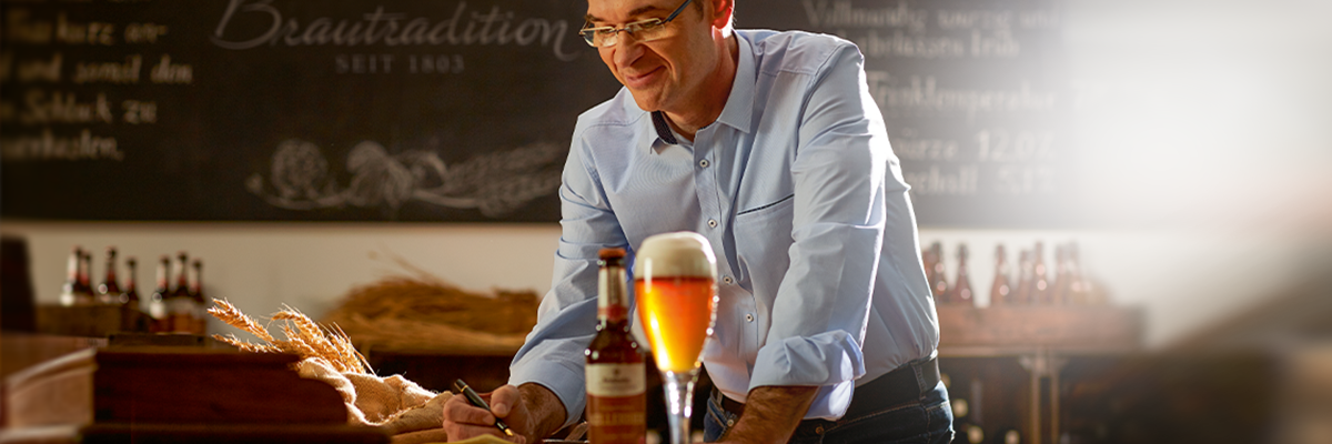 Ein Mann in einem hellblauen Hemd steht an einer Theke und schreibt auf einem Notizblock. Vor ihm steht ein Glas Kellerbier Bier und die dazugehörige Flasche.