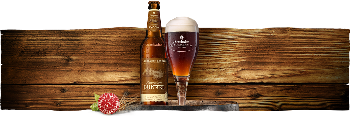 Eine Flasche Krombacher Dunkel Bier steht neben einem Glas mit schaumigem Bier. Im Hintergrund ist eine Holzoberfläche sichtbar.