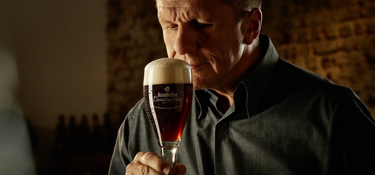 Ein Mann hält ein Glas mit dunklem Bier, das eine cremige Schaumkrone hat. 