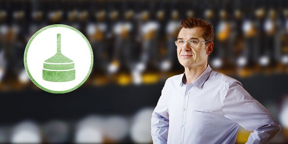 Ein Mann steht mit verschränkten Armen in einem hellen Hemd. Im Hintergrund sind mehrere unscharfe Objekte zu sehen, und links ist ein grünes Symbol eines Trichters.