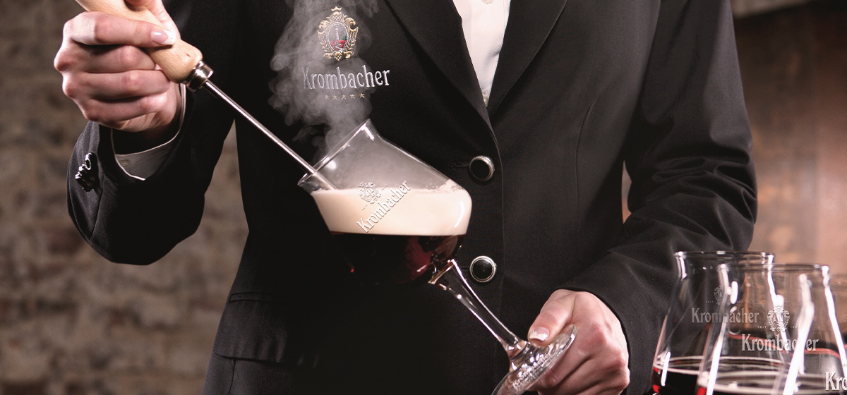 Ein Kellner in einem schwarzen Anzug hält ein Glas mit Dunkelbier und einem Rührgerät. 